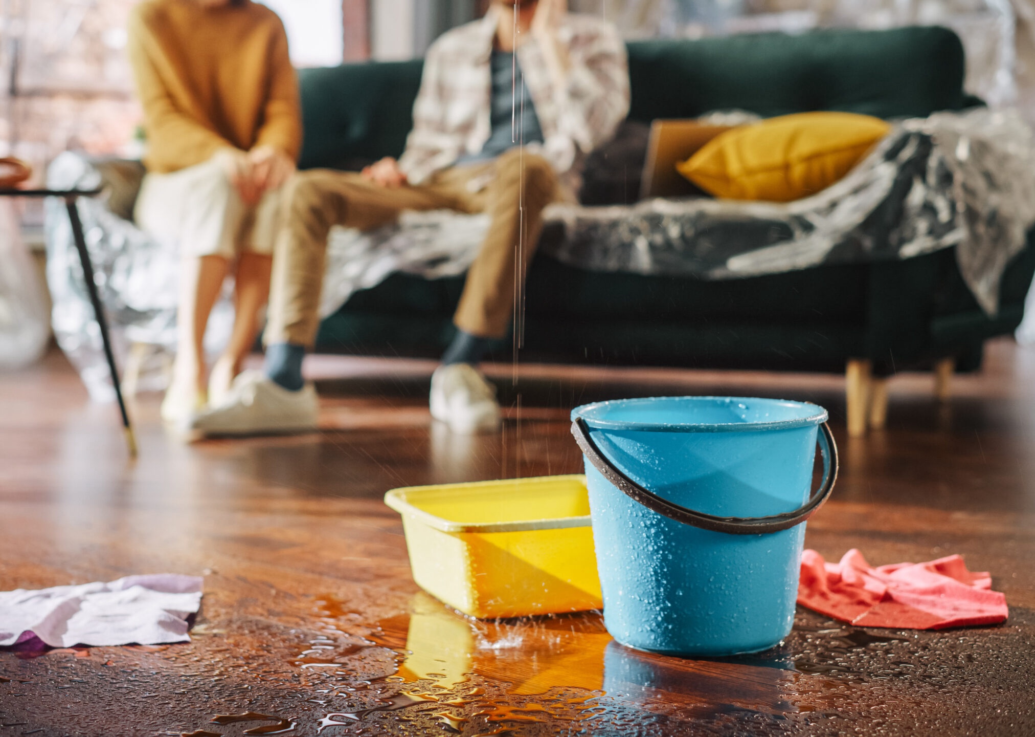 Roof is Leaking, Pipe Rupture at Home: Water Drips into Buckets in Living Room. Angry Couple in Background Calling Insurance Company, Screaming into Phone in Frustration, Trying to Find Plumber Charlotte Water Damage Restoration